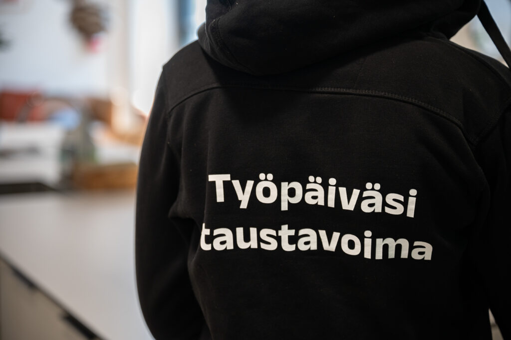 Tässä on kuva meitalaisen selästä ja hänen päällään olevasta hupparista, jossa teksti "työpäiväsi taustavoima"